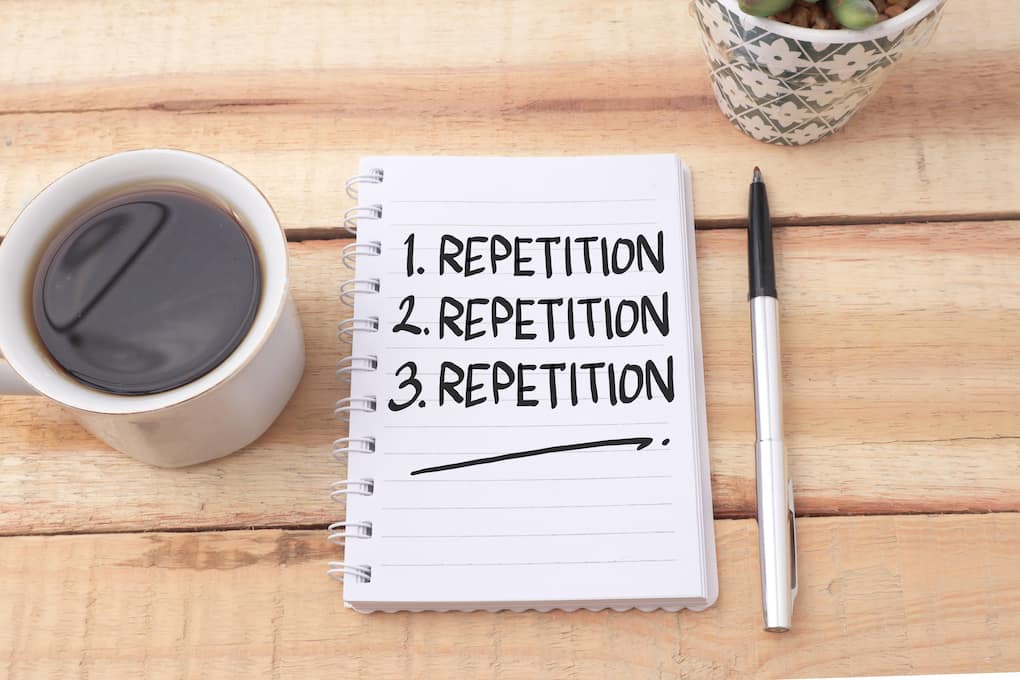 the word repetition written three times on a notepad on a wooden table next to a cup of coffee and a pen