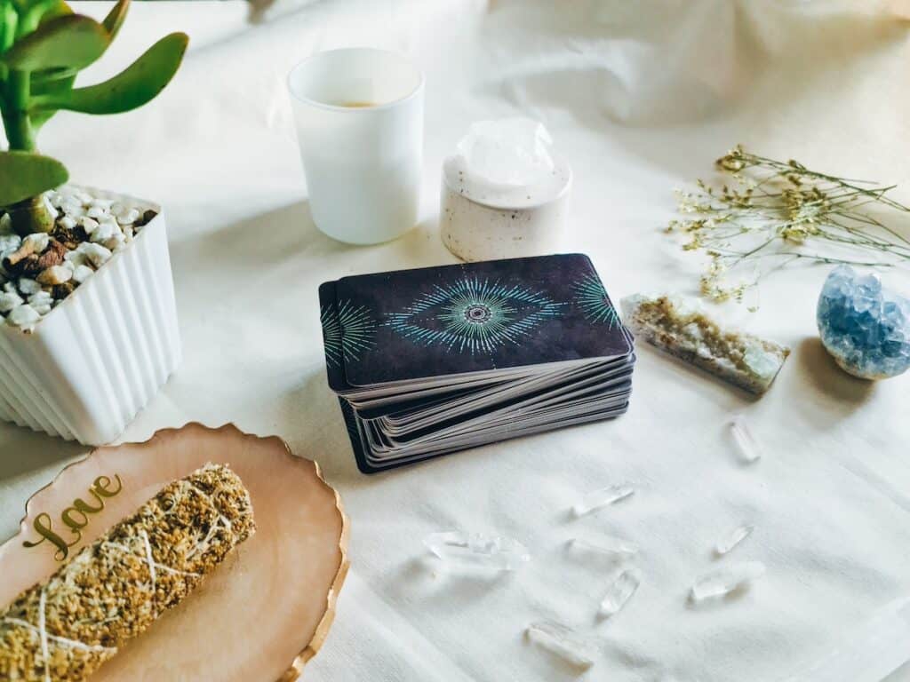 Selection of tarot related objects on white cloth background including tarot card deck, crystals and sage smudge stick