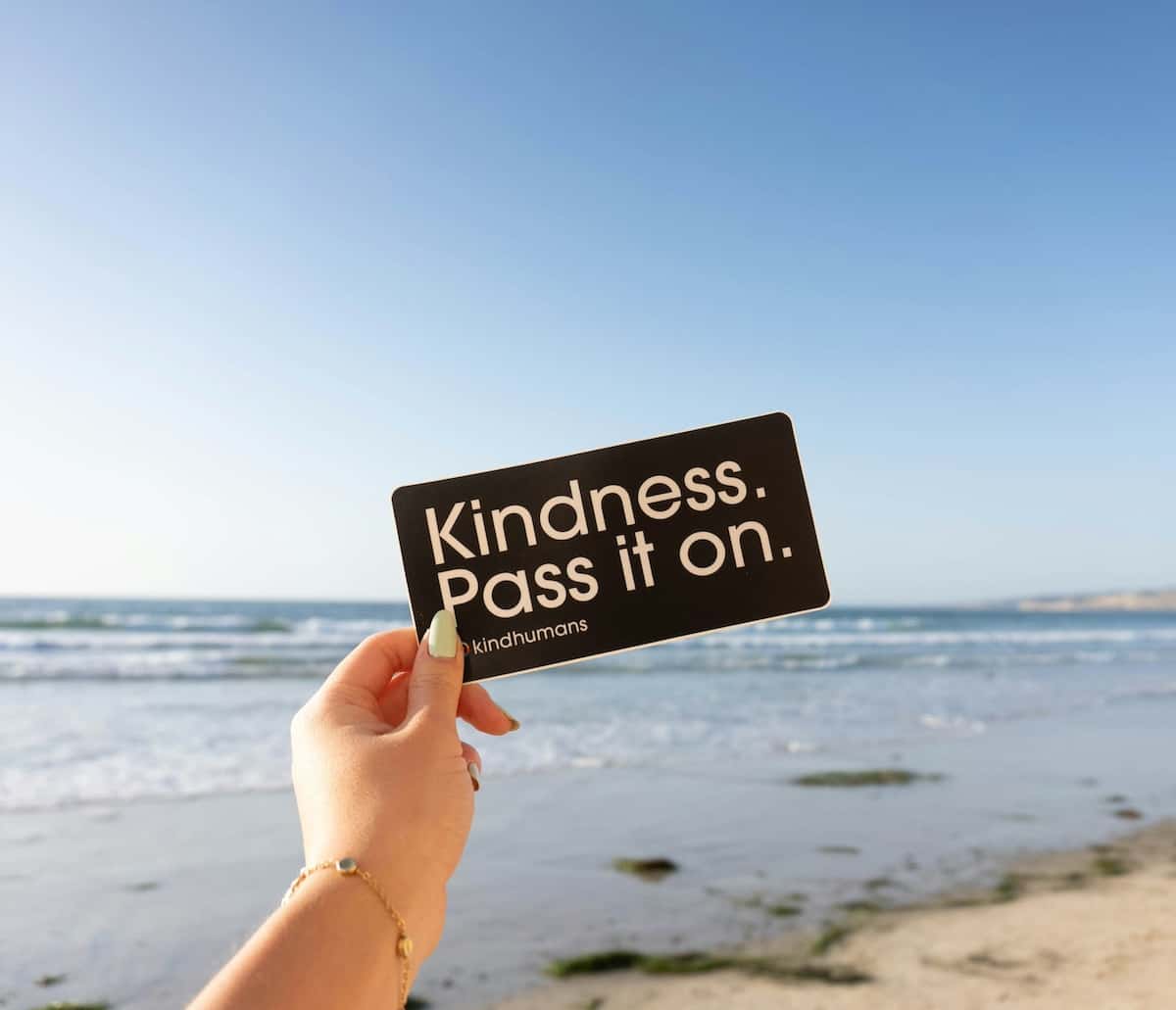 close up of hand holding sign on the beach which reads "kindness. pass it on."