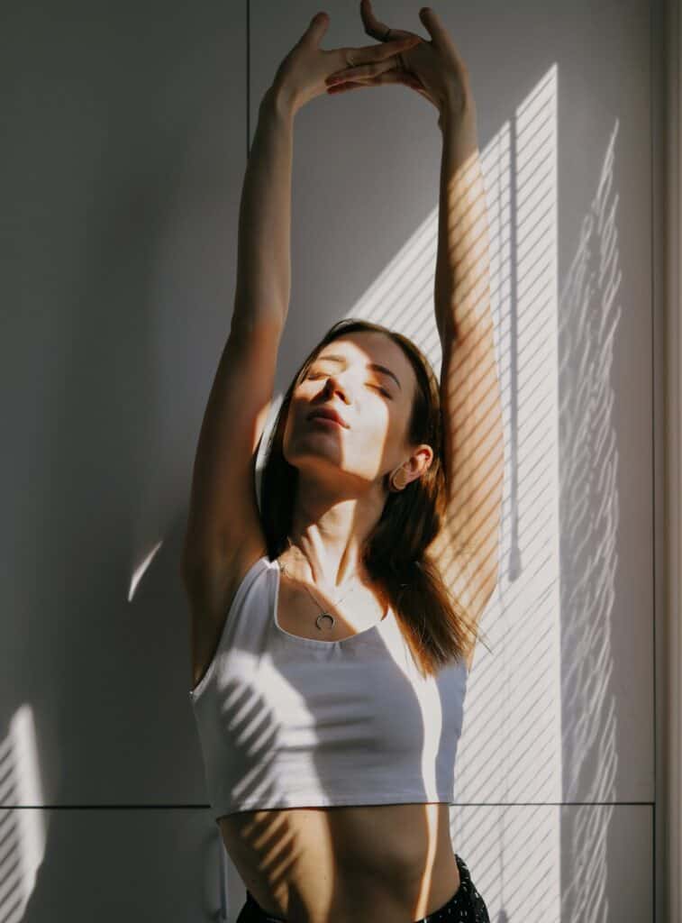 woman looking mindful with eyes closed stretching her arms overhead in the sunlight through her window